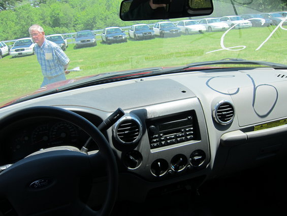 Interior view of a car dashboard with a man standing by several parked cars in the background