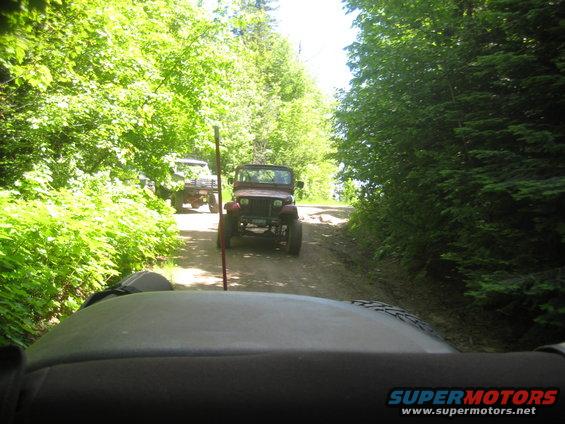 Two Jeep Wranglers driving on a dirt road surrounded by trees. The front Jeep is a darker color, the second is reddish