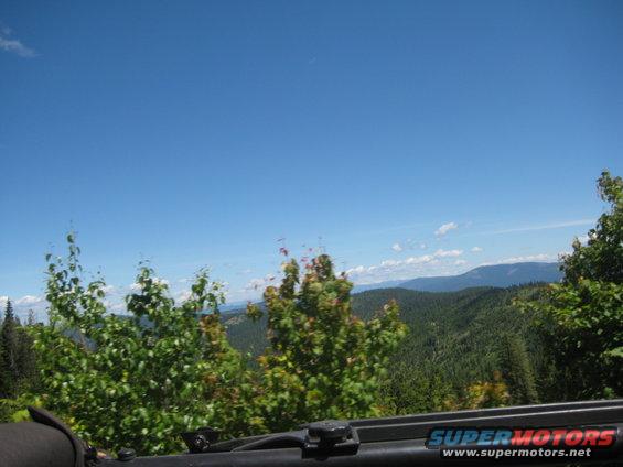 View of forested mountains under a blue sky, seen from inside a vehicle. Watermark reads SUPERMOTORS.NET
