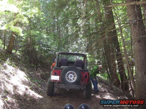 Red Jeep Wrangler on a wooded trail, rear view. License plate is visible