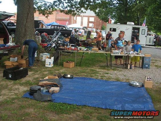 Outdoor swap meet with tables of parts, a motorcycle, and several vehicles in the background