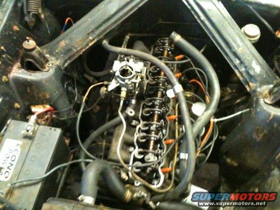 Engine bay view of a V8 engine with carburetor and battery. Some hoses and wiring are visible