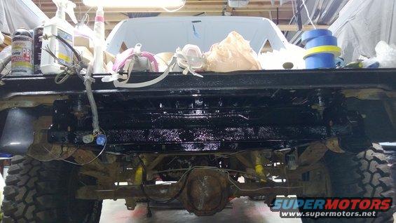 Rear view of a Ford Bronco undergoing work, showing the axle, frame, and suspension components. The bed is visible above, with tools and supplies