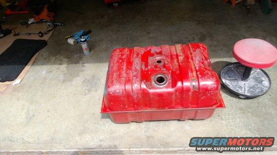 Red fuel tank with rust and peeling paint, sitting on a concrete floor next to a rolling stool