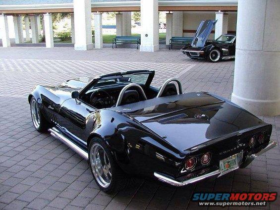 Black convertible Corvette from rear-right angle, chrome side pipes, aftermarket wheels, another Corvette with hood up in background