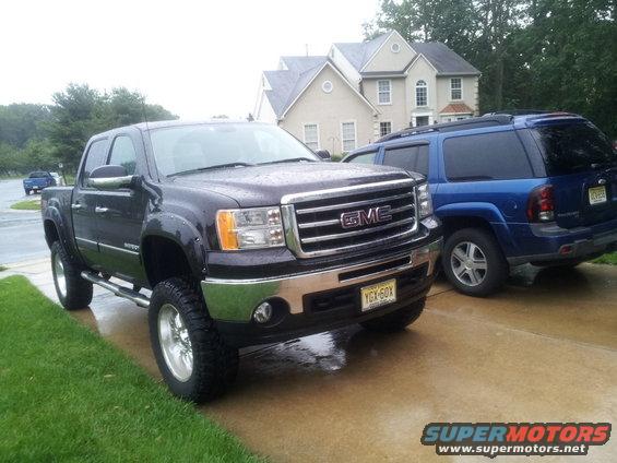 Dark GMC Sierra pickup truck with chrome grille and aftermarket wheels parked on a wet driveway next to a blue SUV