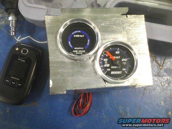 Air/fuel ratio and boost gauges mounted on a metal plate, next to a flip phone