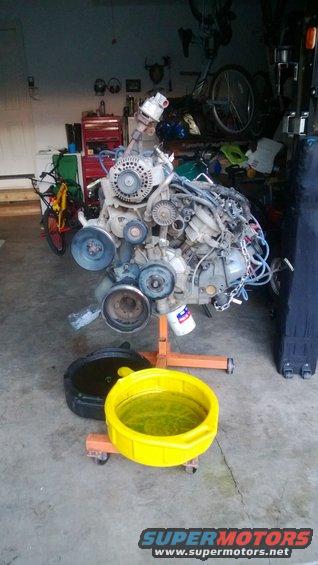 Engine on an orange stand with oil drain pans below. Garage background with tools and bikes