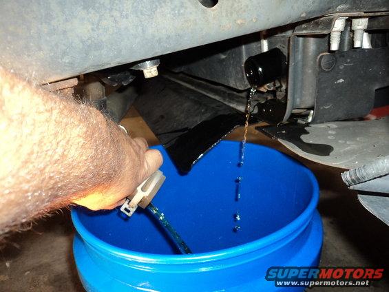 hand holding tool to drain dark fluid into blue bucket under vehicle chassis