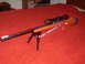 1990 Max II - A rifle equipped with a scope and bipod is displayed on a red fabric surface.