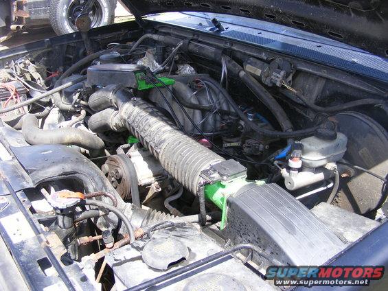 Engine bay with hoses, belts, and air intake system visible. Another vehicle's wheel is in the background