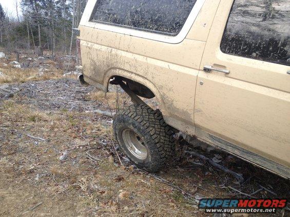 Tan Ford Bronco with mud splatters, seen from the rear quarter panel, off-road tires, in a wooded area