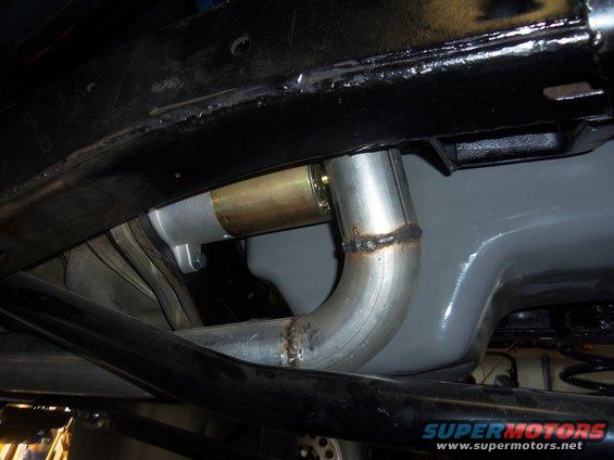 Close-up of a vehicle's undercarriage showing a starter motor connected to an exhaust pipe. Welds are visible on the pipe