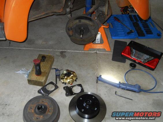 Car brake drum, rotor, caliper, and tools laid out on a garage floor