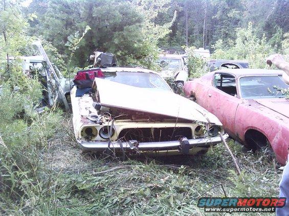 Yellow 1970s muscle car with damaged front end and hood, parked in a junkyard