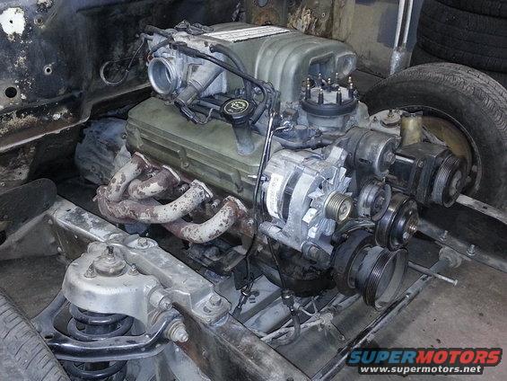 Engine assembly with exhaust manifolds, alternator, and pulleys. Chassis components visible