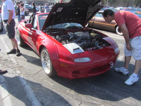 Red Mazda Miata with aftermarket widebody kit and hood open, showing engine. Man in red shirt inspects engine. Other cars in background