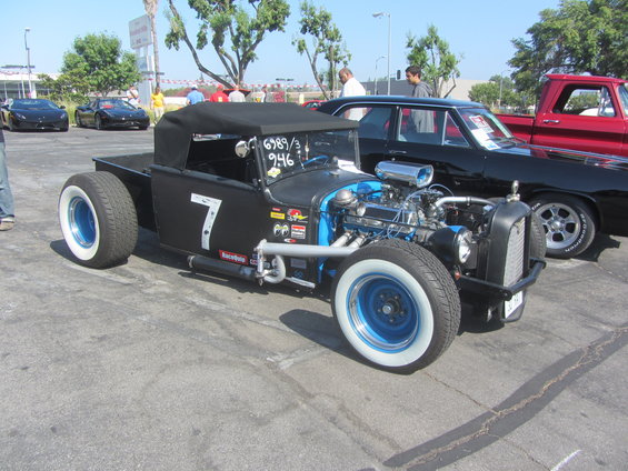 Black hot rod pickup with number 7 on door, exposed engine, and whitewall tires. Other cars visible in background