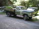 1979 Ford Bronco - A green flatbed truck and a dark SUV are parked on a gravel road in front of a wooden cabin.