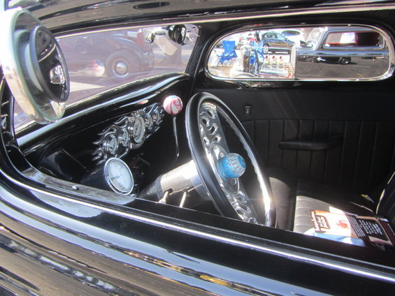 Interior of a black hot rod with custom gauges, steering wheel, and a magazine on the seat. Another car is visible in the side mirror