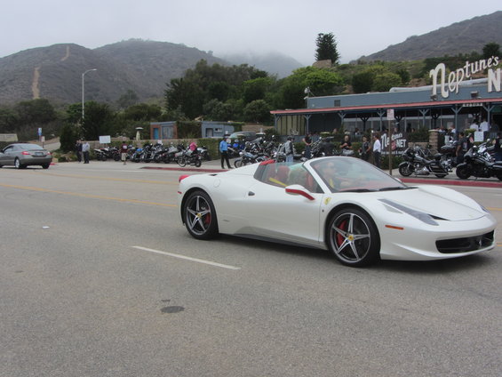 White convertible sports car driving on a road, with a restaurant and many motorcycles parked in the background