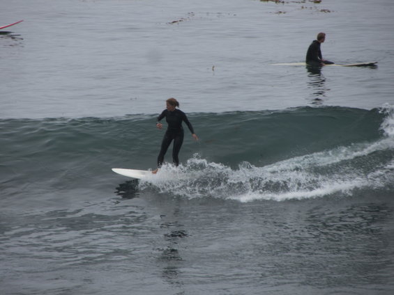 Surfer in a black wetsuit rides a wave on a white surfboard, with another surfer in the background