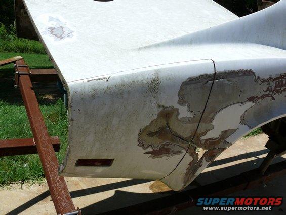 White car body panel, possibly a fender or hood, mounted on a metal stand. Paint is chipped and rusted in areas