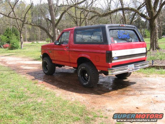 Red 1992 Ford Bronco with a black removable top, lifted suspension, and aftermarket wheels parked on a dirt path