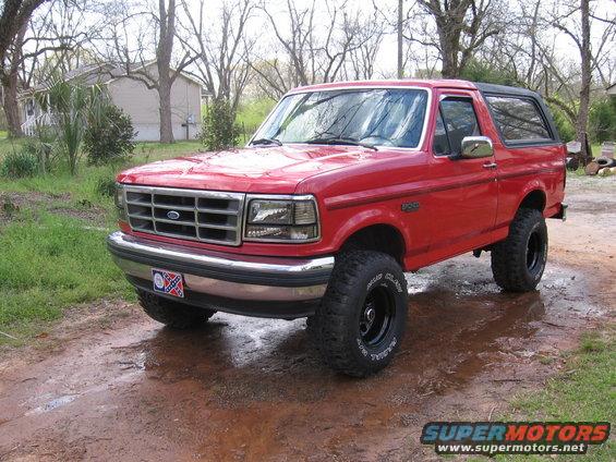 Red 1992 Ford Bronco parked on a muddy dirt road with trees and a house in the background