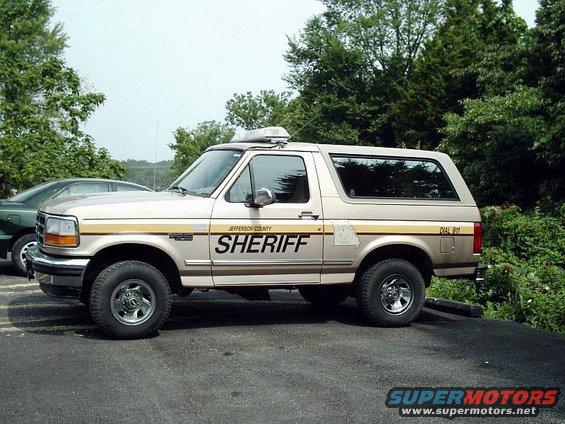 Side view of a tan Ford Bronco with 'SHERIFF' on the door and a light bar on the roof