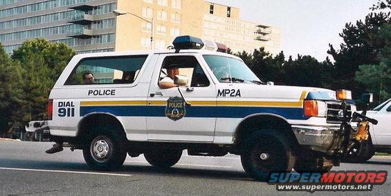 White 1992 Ford Bronco police SUV with blue and yellow stripes, marked 'POLICE' and 'MP2A', driving on a road