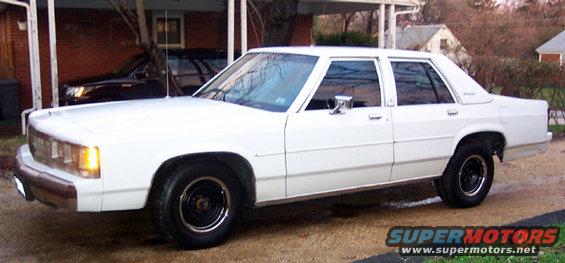 White sedan parked on a gravel driveway, with another dark vehicle in the background under a carport