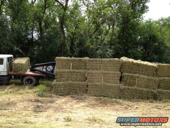 Flatbed truck carrying a hay bale, parked next to a large stack of hay bales