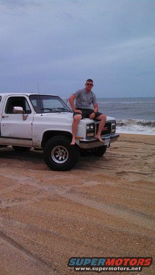 Man sitting on the hood of a white GMC pickup truck parked on a sandy beach with the ocean in the background