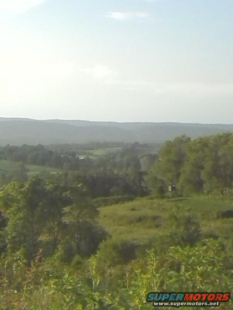 Rural landscape with rolling hills, trees, and vegetation under a hazy sky. Watermark reads SUPERMOTORS.NET
