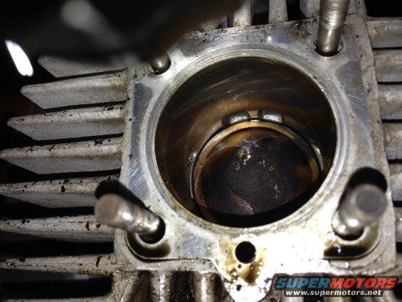 Motorcycle engine cylinder with piston visible inside, showing carbon buildup. Fins on the exterior are visible