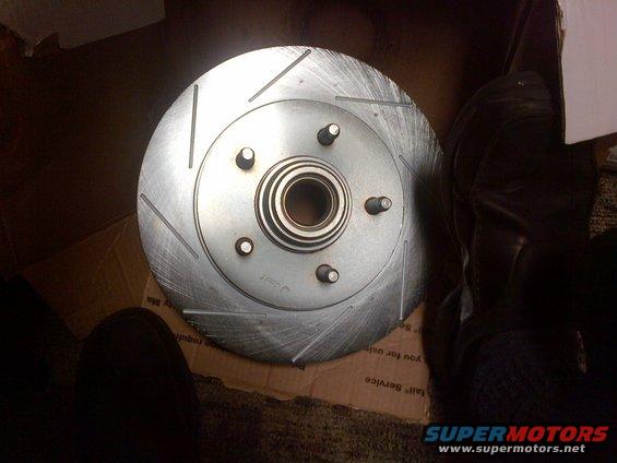 Slotted brake rotor with hub and five lug nuts, resting on cardboard