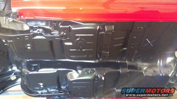 Underside of a red vehicle with black undercoating, showing frame rails and floor pan