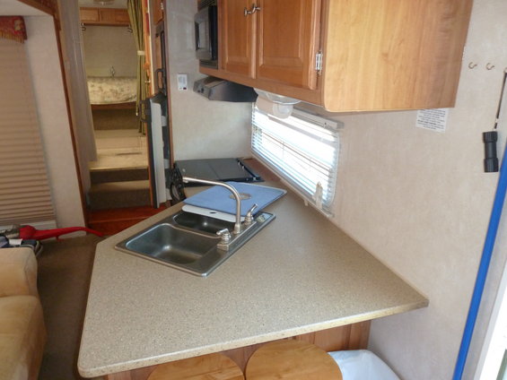 Camper interior with a sink, countertop, and cabinets. A bed is visible in the background