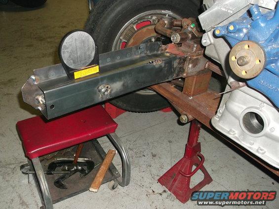 Close-up of vehicle frame with engine parts and a leveling gauge. A red stool and jack stands are visible