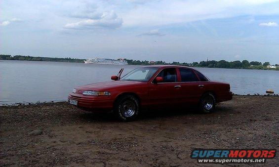 Red 1992 Ford Crown Victoria sedan with driver's door open, parked on a dirt bank by a river