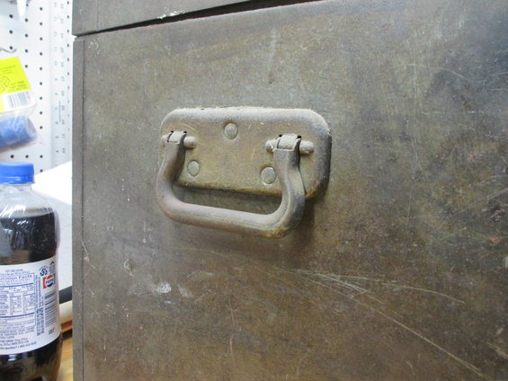 Metal trunk handle on a weathered, scratched metal surface. A bottle of dark liquid with a blue cap is visible to the left
