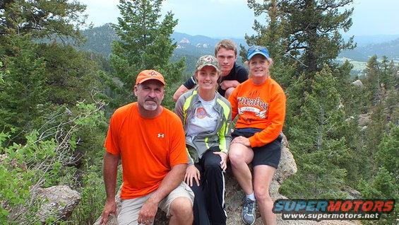 Four people in casual outdoor clothing sit on rocks in a mountainous, wooded area. A watermark reads SUPERMOTORS