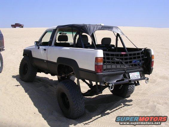 Lifted white and black pickup truck with a soft top, modified for off-road use, parked on sand. Another red pickup is visible in the distance