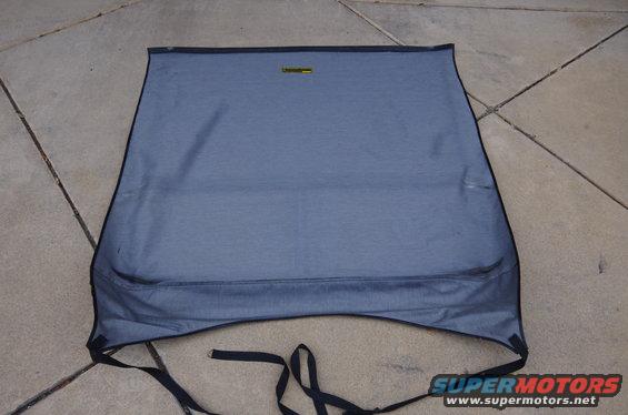 Blue fabric convertible top with black trim and straps, laid out on a concrete surface. A yellow tag is visible