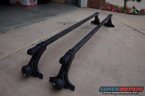 Pair of black roof rack crossbars with adjustable mounts, resting on a concrete driveway