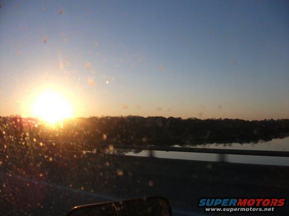 Sunset over a body of water with trees on the horizon, seen through a dirty windshield