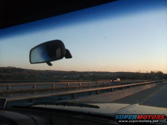 View from inside a vehicle looking out the windshield at a highway overpass and distant hills at sunset. A side mirror is visible