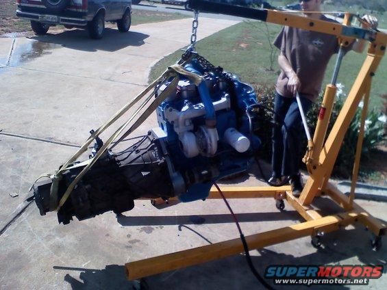 Engine and transmission assembly suspended from an engine hoist, with a Ford Bronco in the background