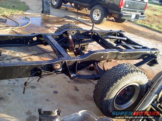 vehicle frame with suspension components and tires, black pickup truck in background